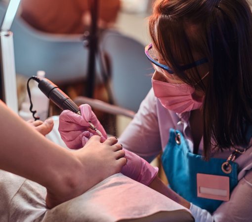 Young cheerful nail master is working on client's toe nails at her pedicure studio.